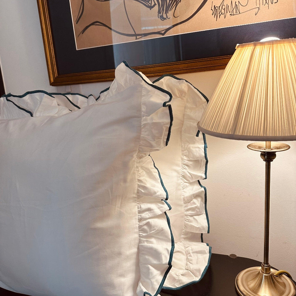 White Pillowcase with Teal trim on a bed next to a lamp with a gold base.