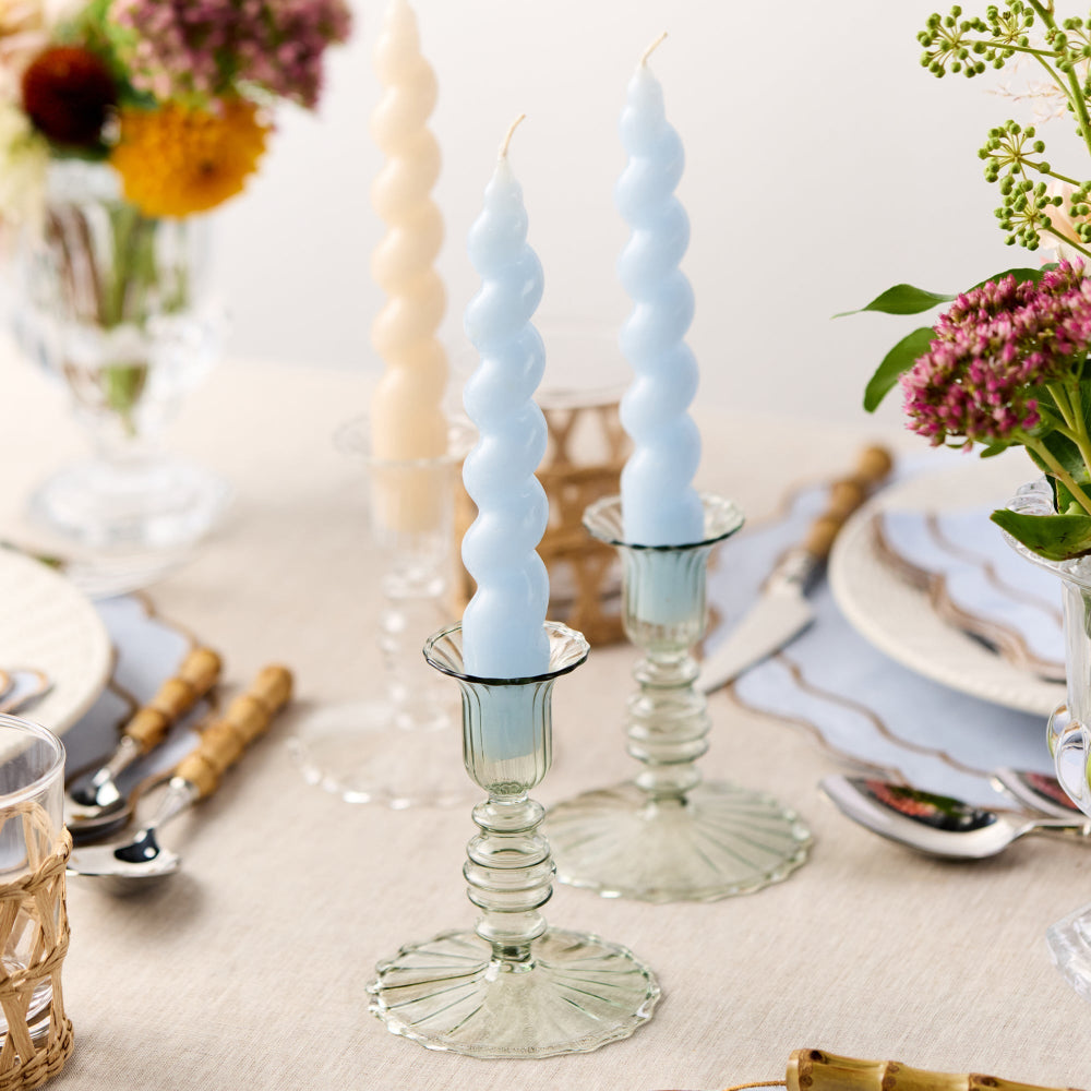 Two spiral-patterned candles in glass candlesticks on a table setting with flowers and cutlery.