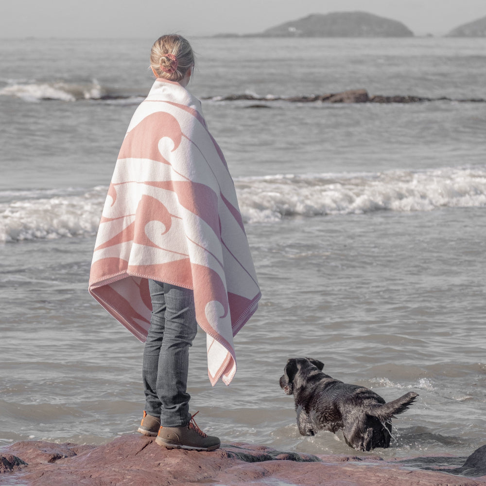 Person wrapped in a pink and white patterned blanket standing on a beach with a dog.