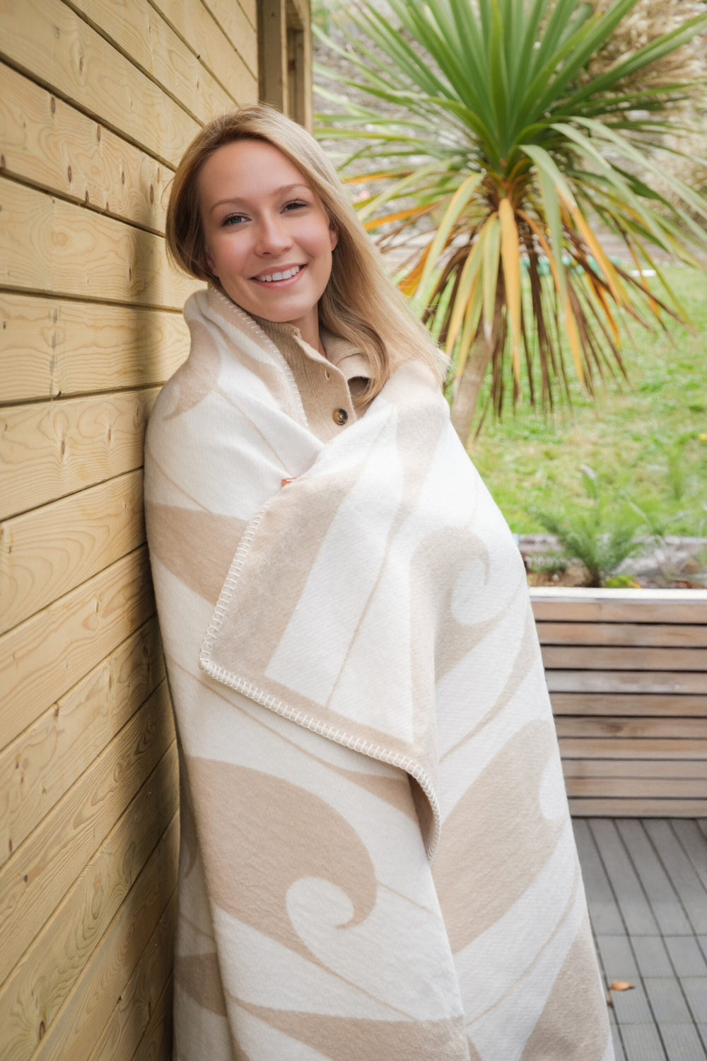 Woman wrapped in a patterned blanket standing against a wooden wall with plants in the background