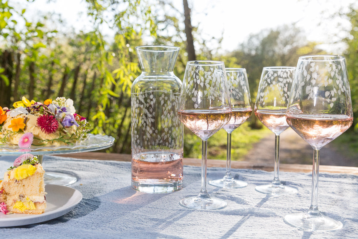 Wine glasses with rose wine on a table outdoors, with a carafe and cake in the background.