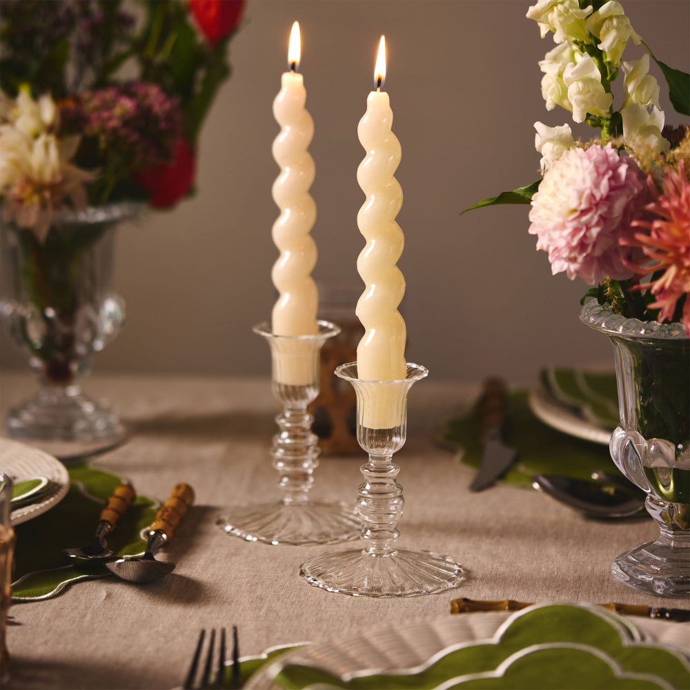 Two spiral candles in glass holders on a table with floral arrangements.