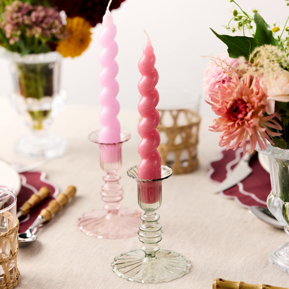 Two pink spiral candles in glass candlesticks on a table with floral arrangements and cutlery.