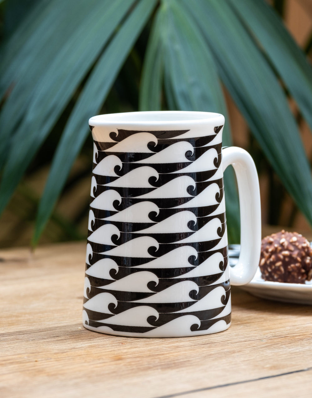 White mug with black wave pattern on a wooden surface with greenery in the background