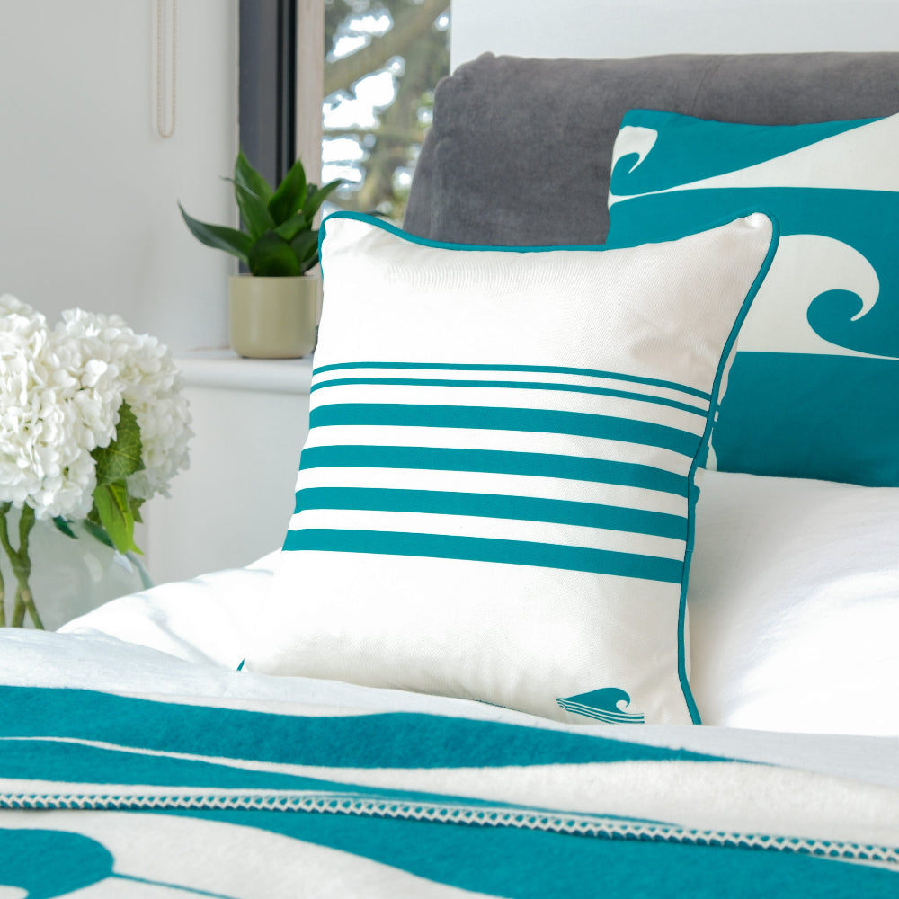 Teal and white striped pillow on a bed with a grey headboard with white flowers in a glass vase on the bedside.