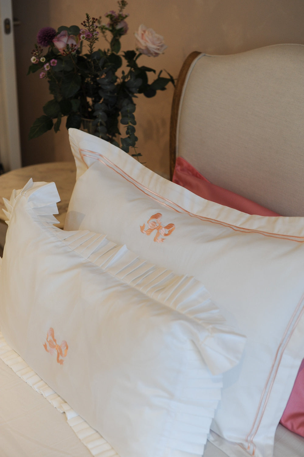 White embroidered pillowcases with bow embroidery and floral arrangement on a beige side table.
