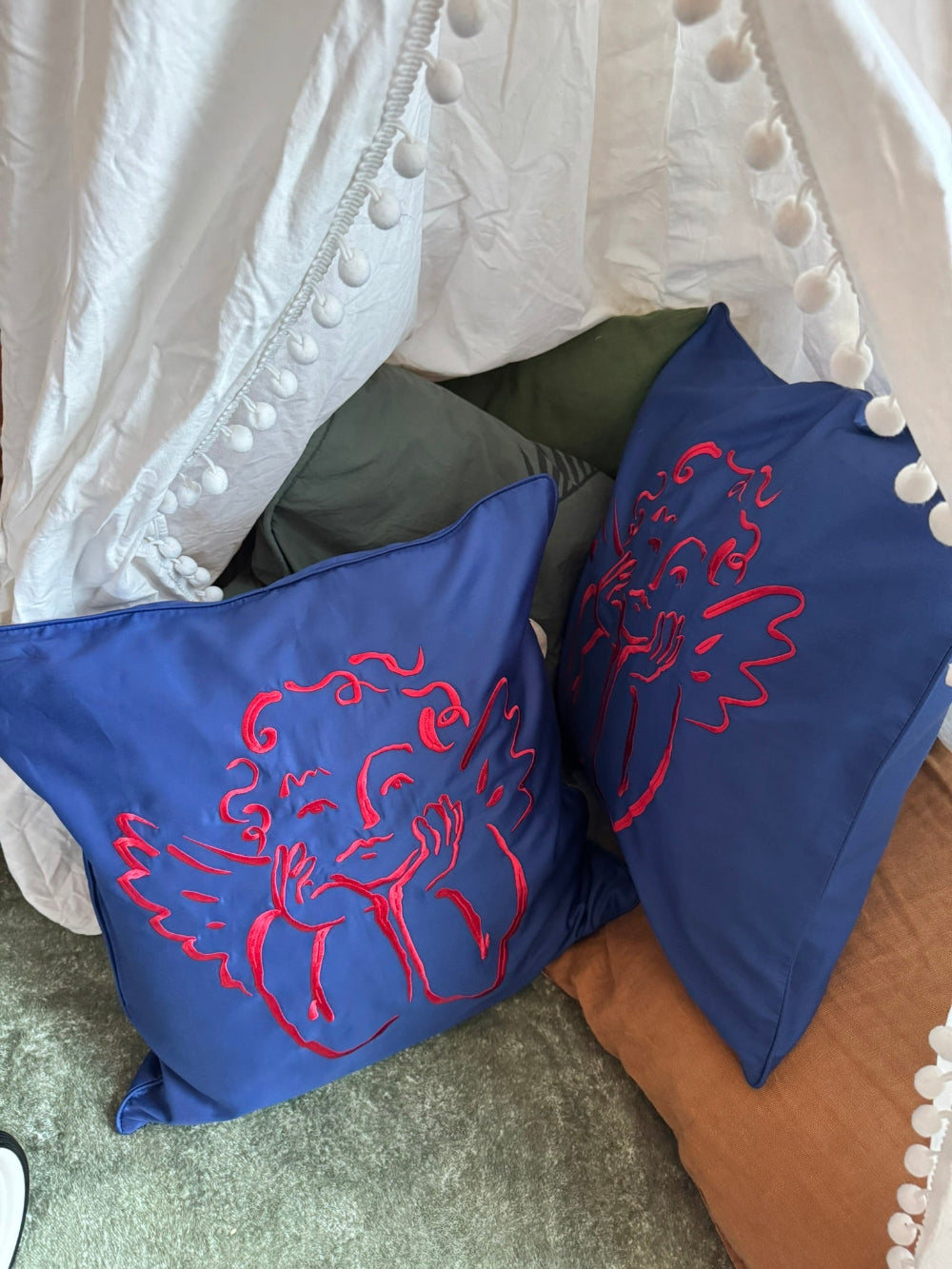 Two blue pillows by Sukun with red cherub patterns on a a floor in a homemade tent.