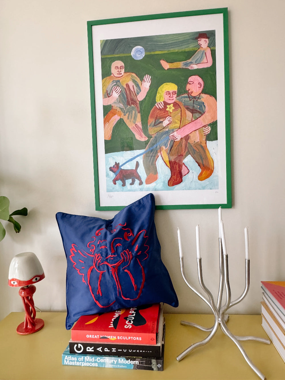 Framed artwork on a wall above books and decorative items with Sukun Blue cushion with red embroidered cherub design.