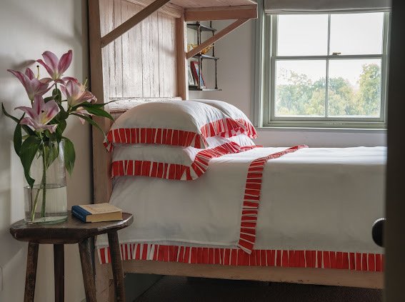 Bed with red and white striped bedding in a room with a window and small table with flowers and books.
