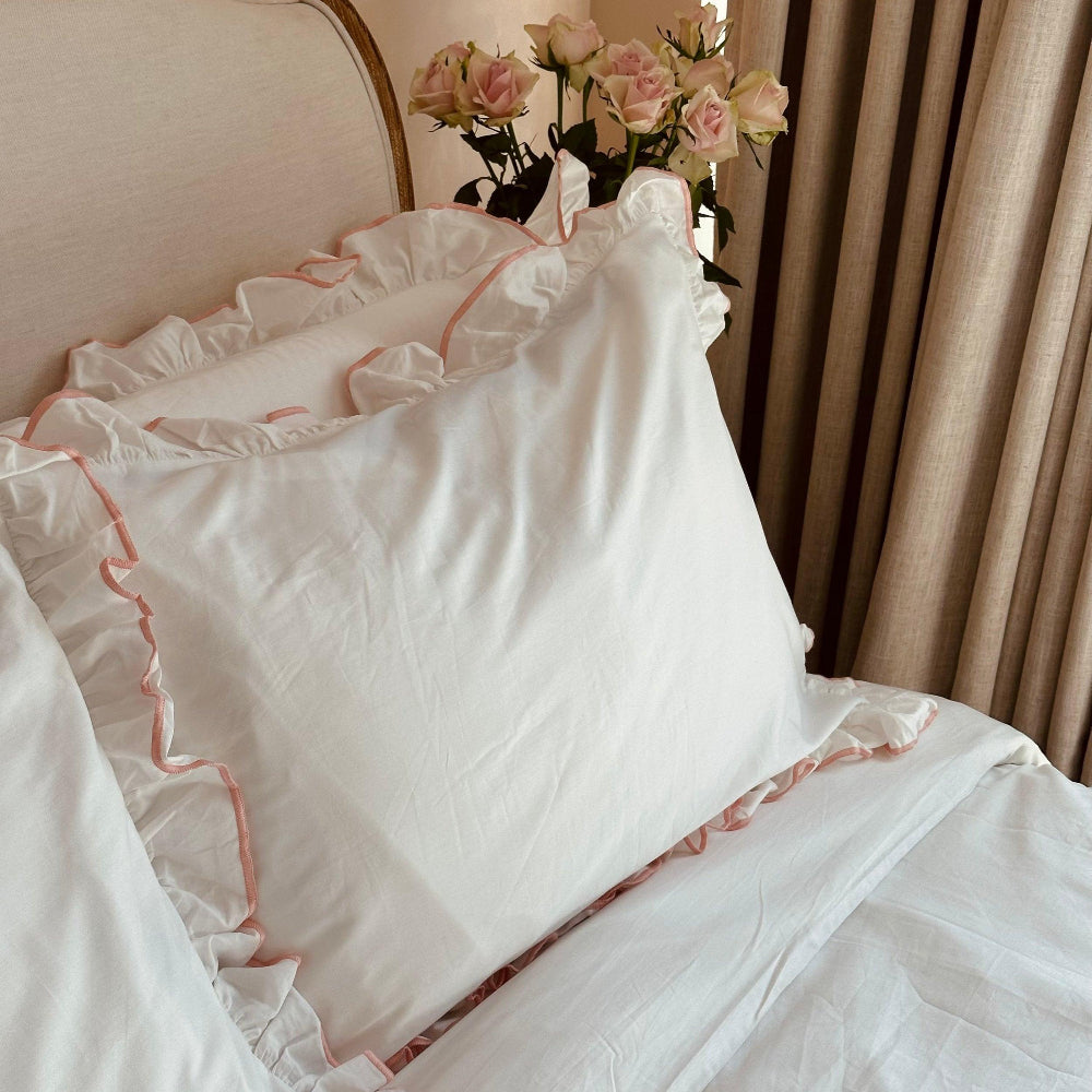 White ruffled bedspread with pink trim on a bed, accompanied by a vase of pink flowers.