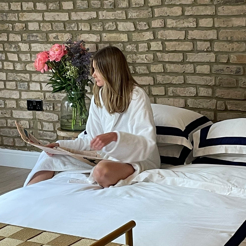 Woman in a white robe reading a newspaper on a bed with a brick wall background