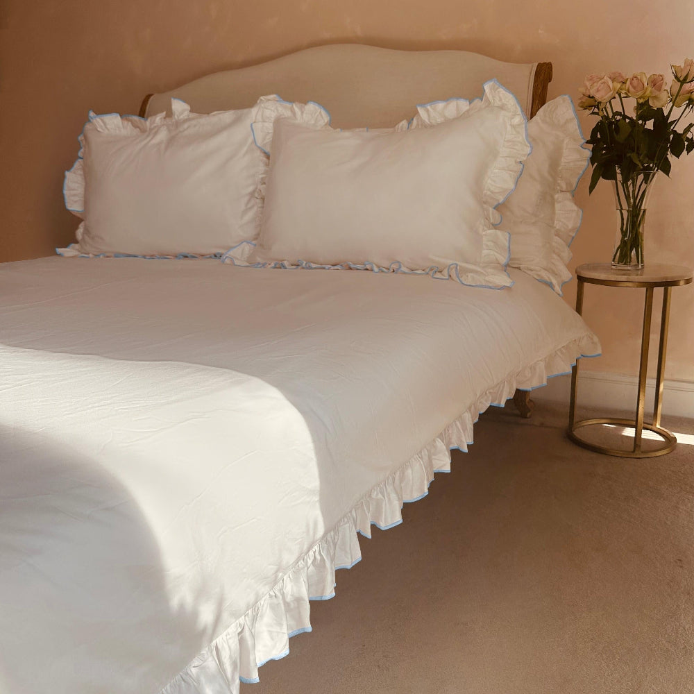 Neatly made bed with white bedding and ruffled edges, next to a small table with flowers.