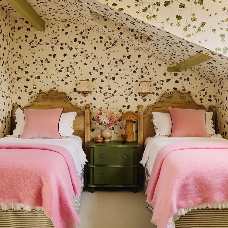 Two twin beds with pink bedding in a room with floral wallpaper and a green nightstand.