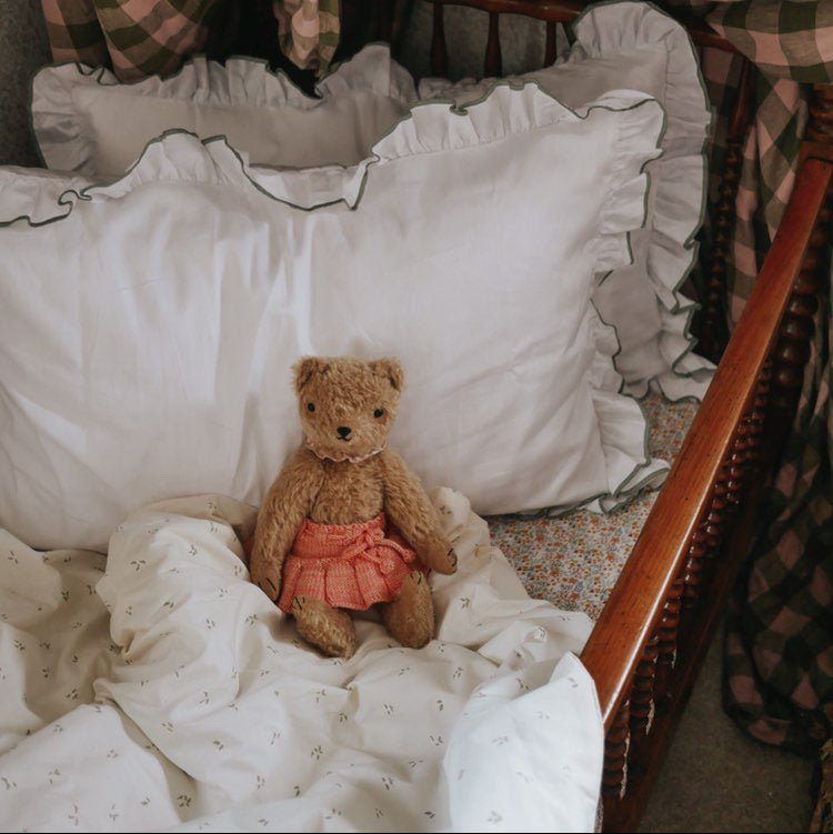 Teddy bear in a crib with white bedding and Olive coloured edges