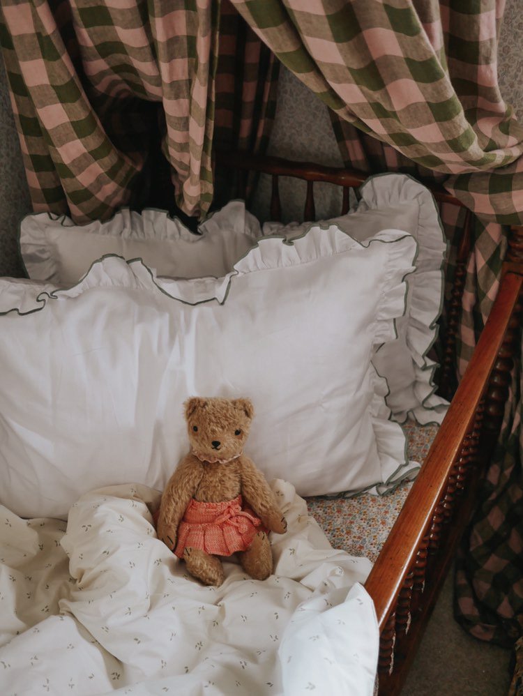 Teddy bear in a pink dress sitting on a white pillow with plaid curtains in the background