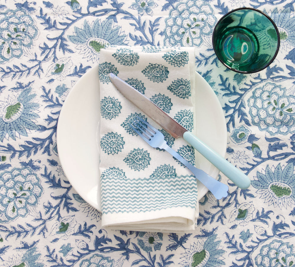 Table setting with a patterned napkin, fork, knife, and glass on a floral tablecloth.