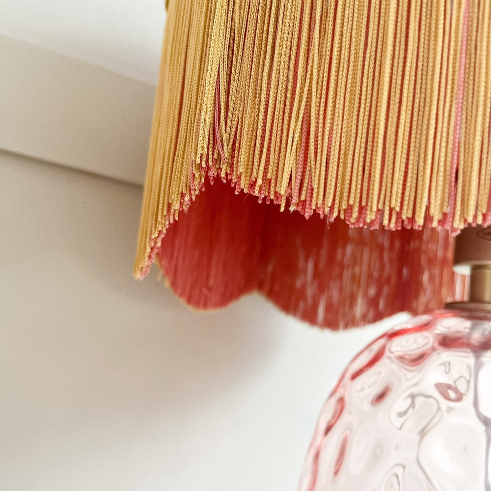 Close-up of a decorative lamp with red fabric and gold fringe against a neutral background
