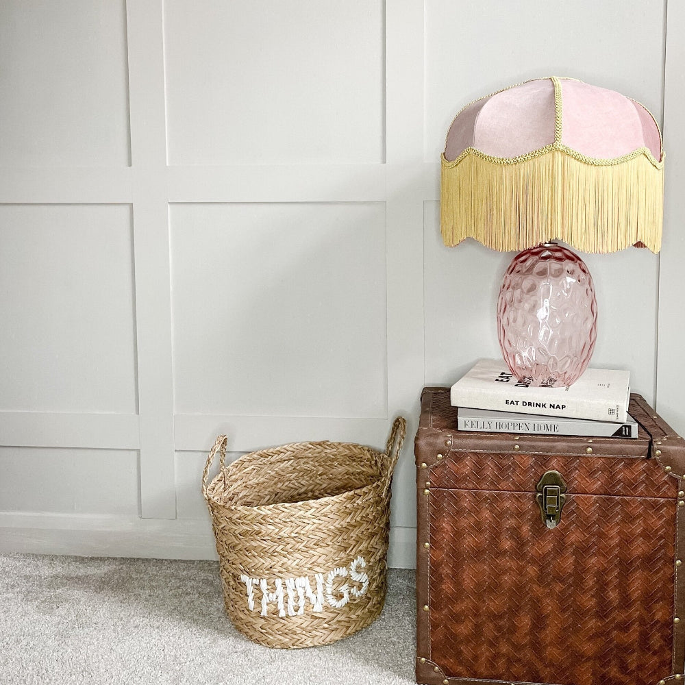 Decorative lamp with pink shade on a wooden chest next to a woven basket with 'THINGS' text.