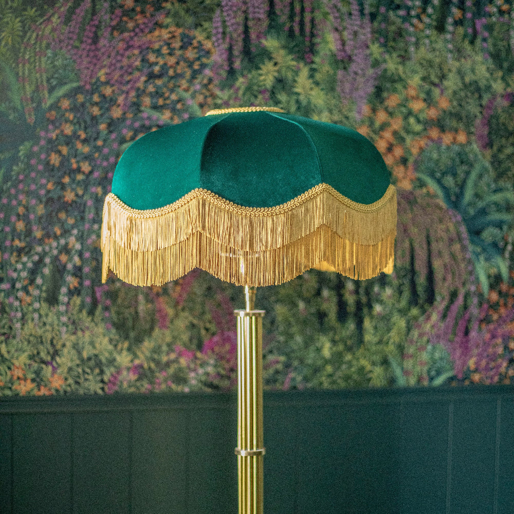 Green lampshade with gold fringe on a stand against a floral wallpapered wall.
