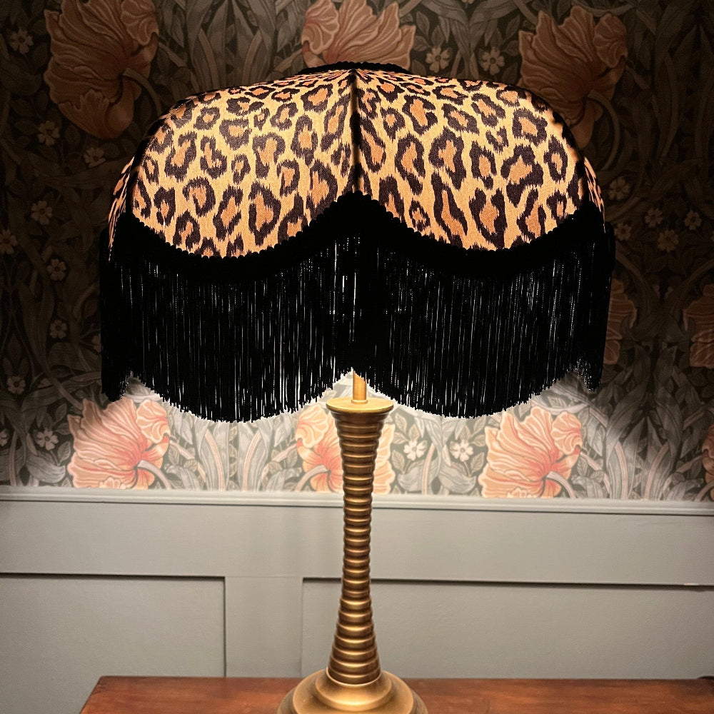 Lamp with leopard print shade and black fringes on a floral wallpapered wall.