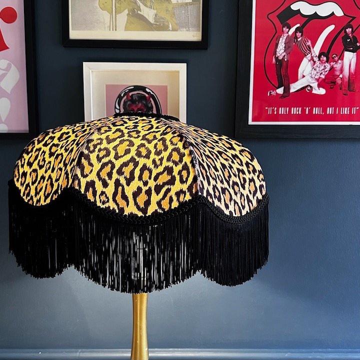 Leopard print lampshade with black fringes on a stand against a dark blue wall with framed posters.
