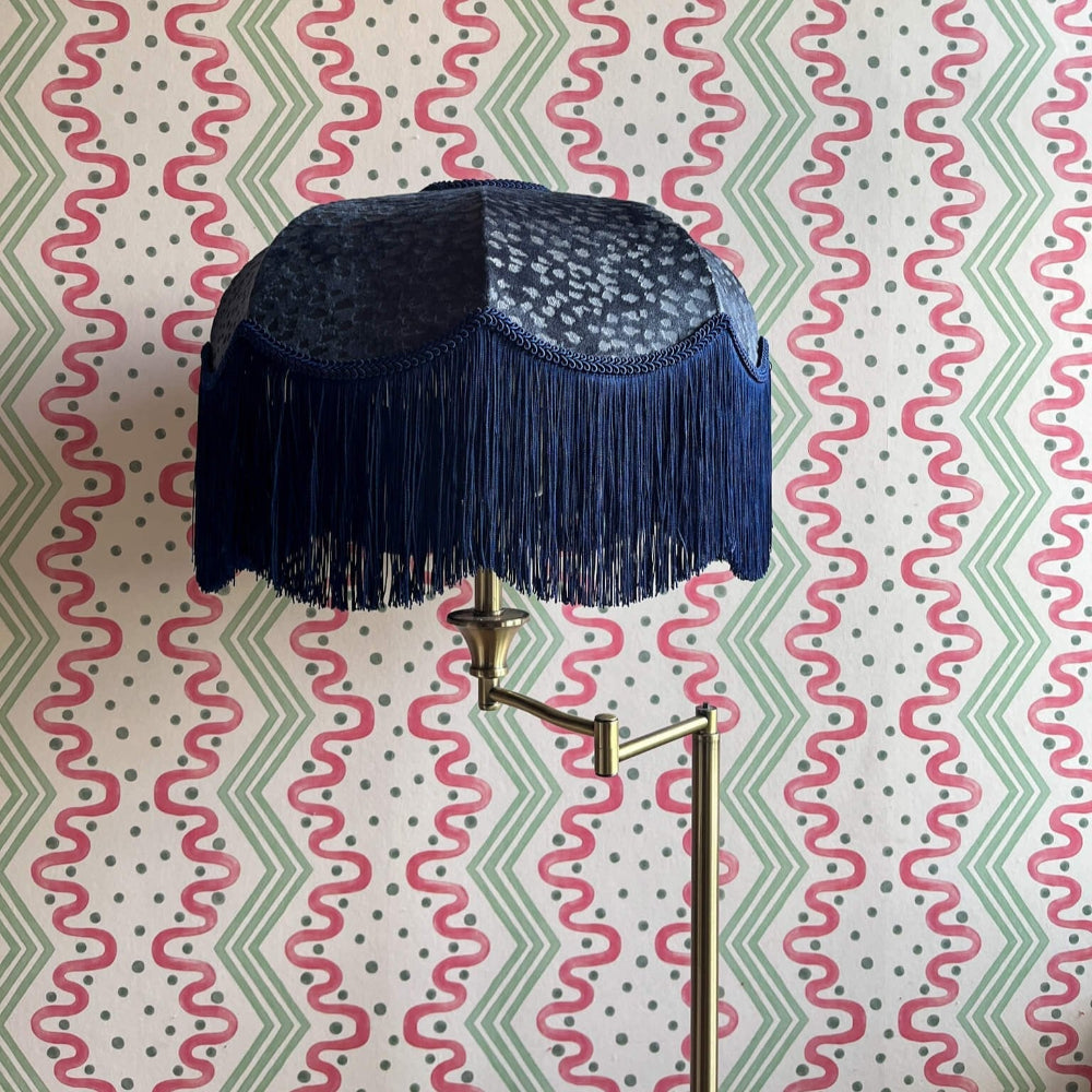 Navy blue lampshade with fringe on a gold arm against a patterned wall.