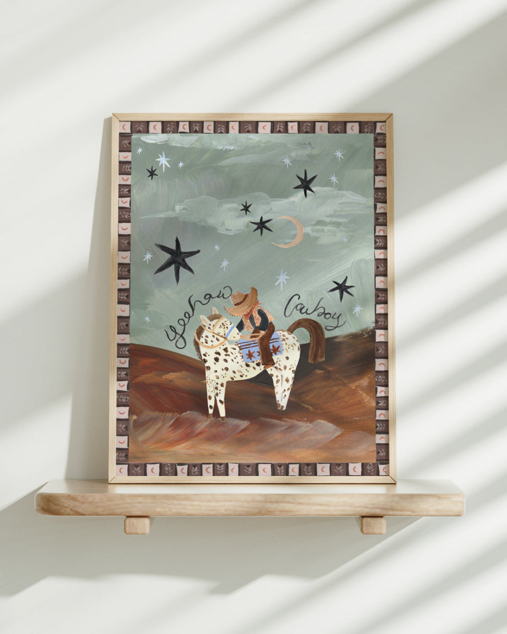 Framed artwork of a cowboy on a horse with stars and moon in the background, displayed on a shelf.
