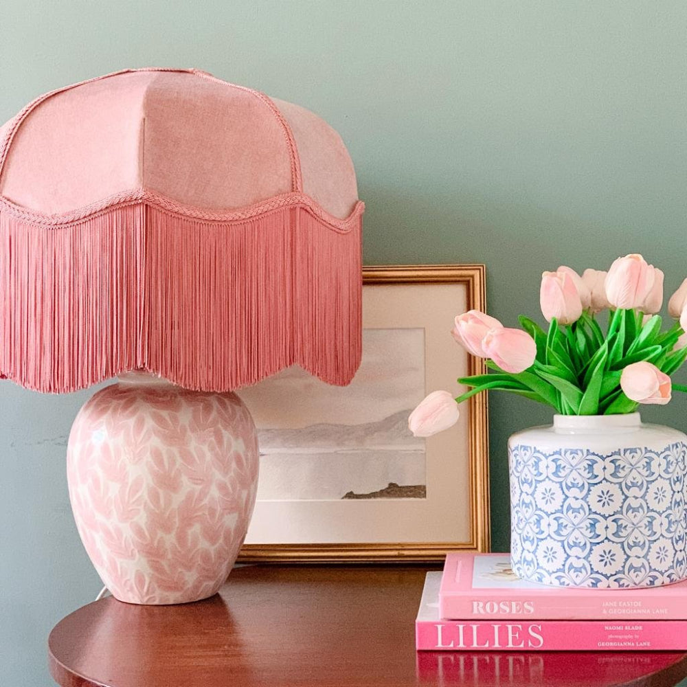 Pink lamp with fringe shade on a table next to a vase of flowers and books.