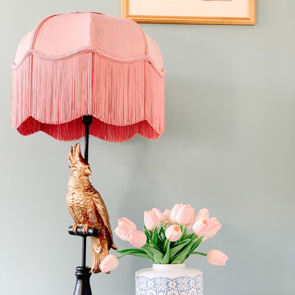 Pink fringed lamp with a gold bird sculpture on a gray wall background