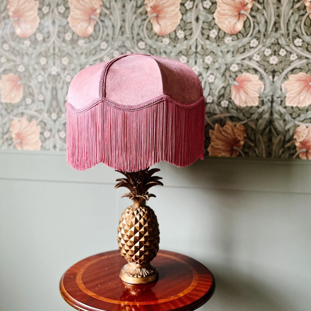Decorative lamp with pink fringed shade and pineapple base against a floral wallpapered wall.