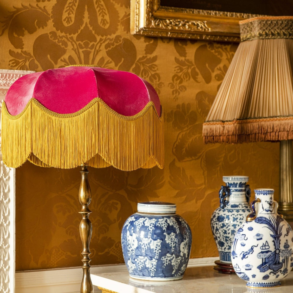 Decorative lamp with pink shade and gold fringe against a patterned wall.
