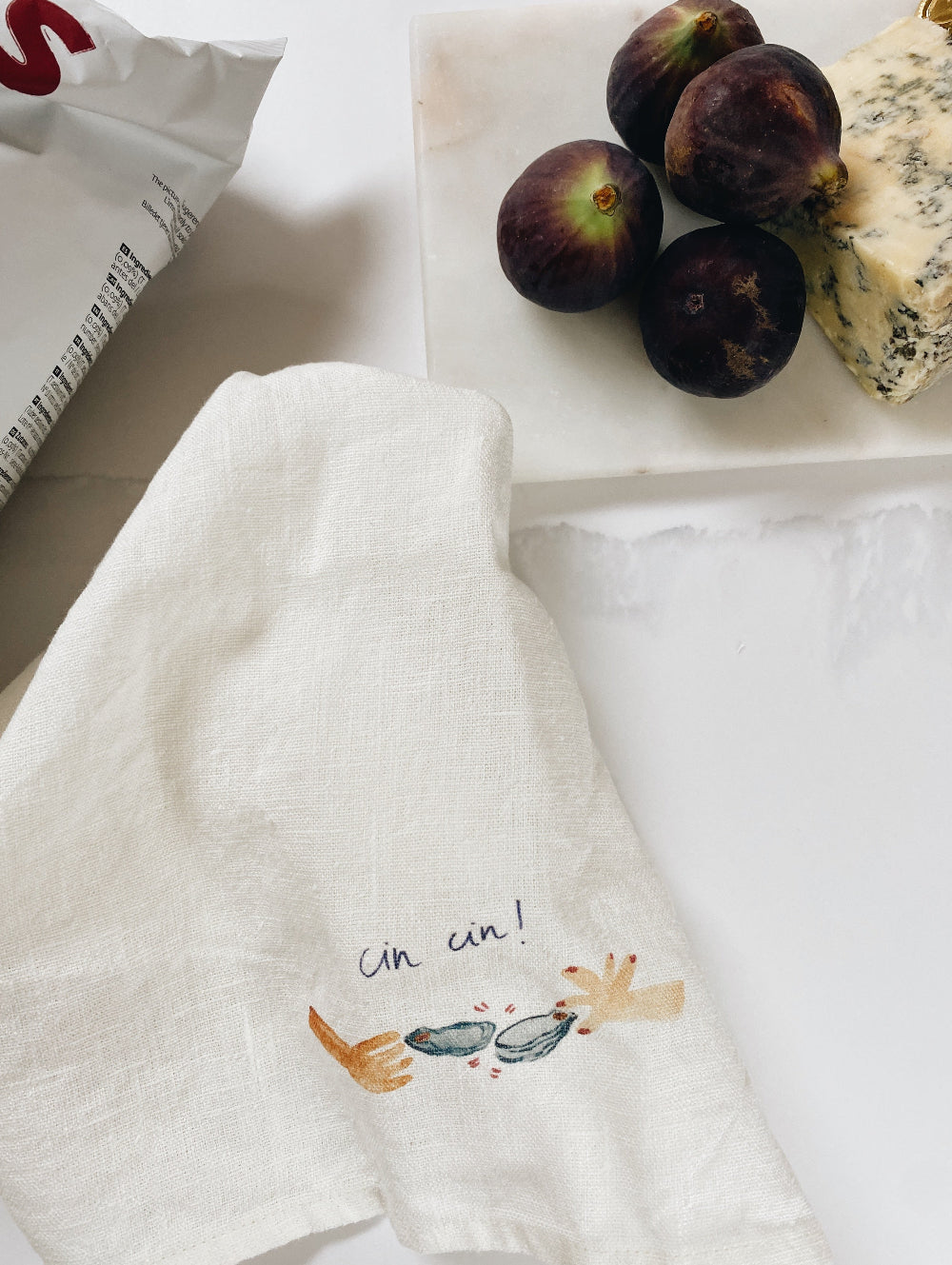 White napkin with 'cin cin!' and wine glass design, placed on a surface with figs and cheese.