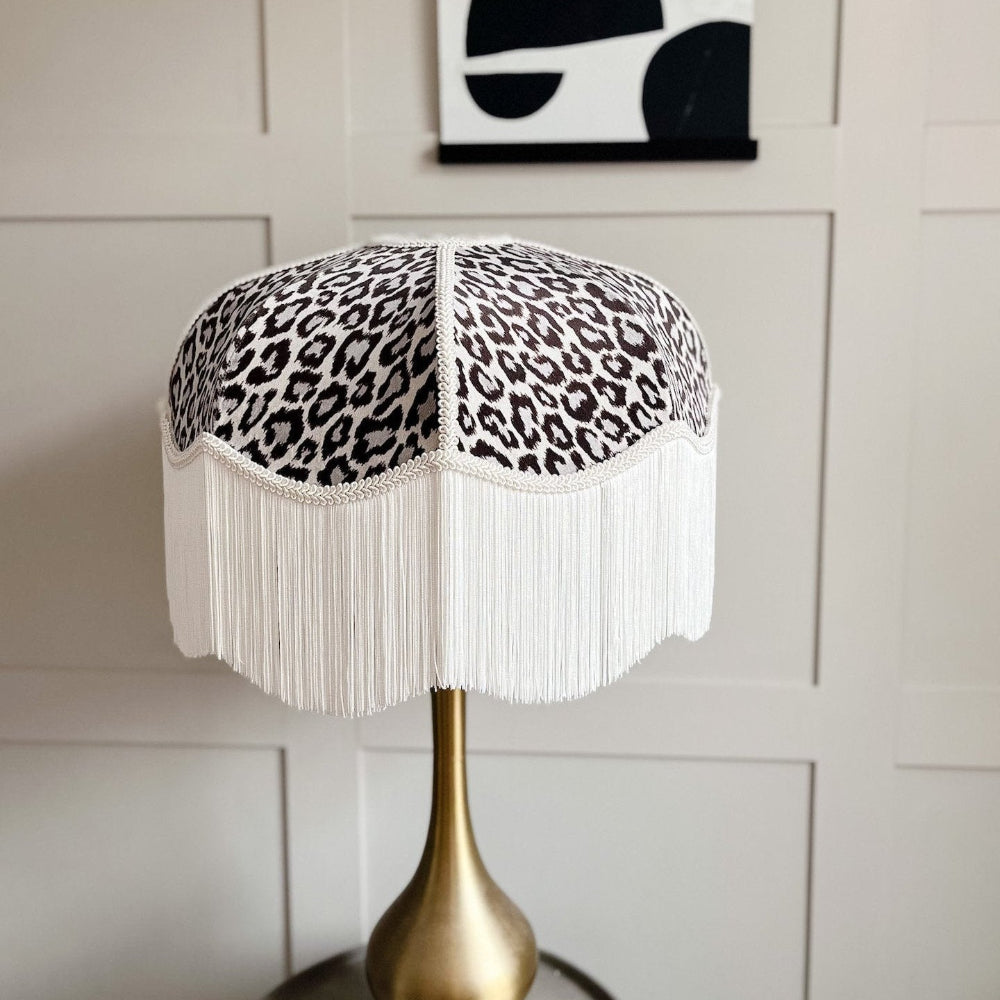 Table lamp with a leopard print shade and white fringe against a neutral wall.