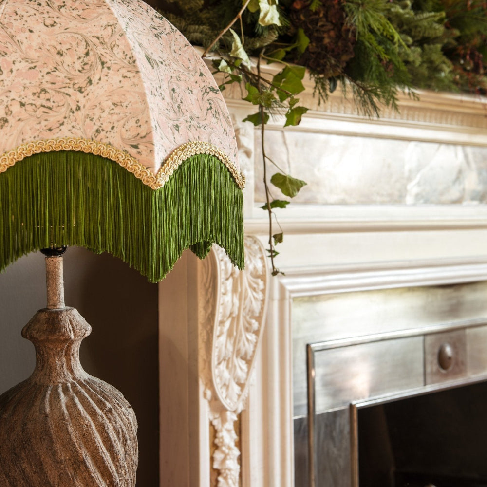 Decorative lamp with a William Morris shade and green fringe in front of a fireplace.