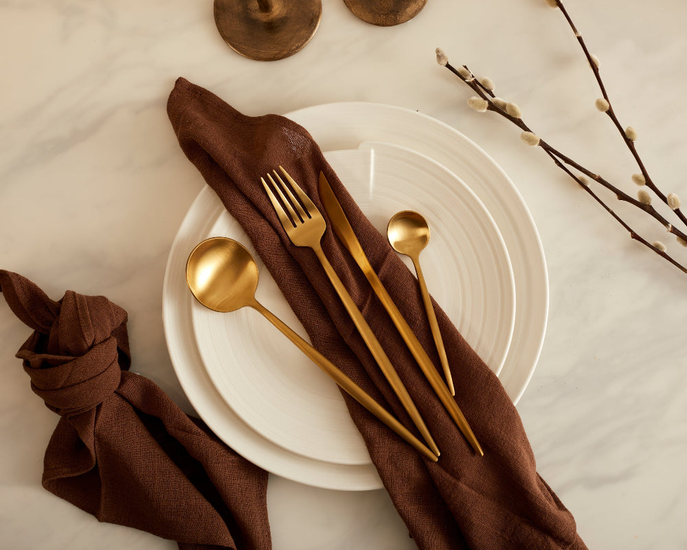 Table setting with gold cutlery and a brown napkin on white plates.