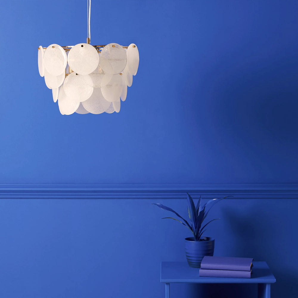 White scalloped-edge pendant light against a blue wall with a plant and books on a table below.