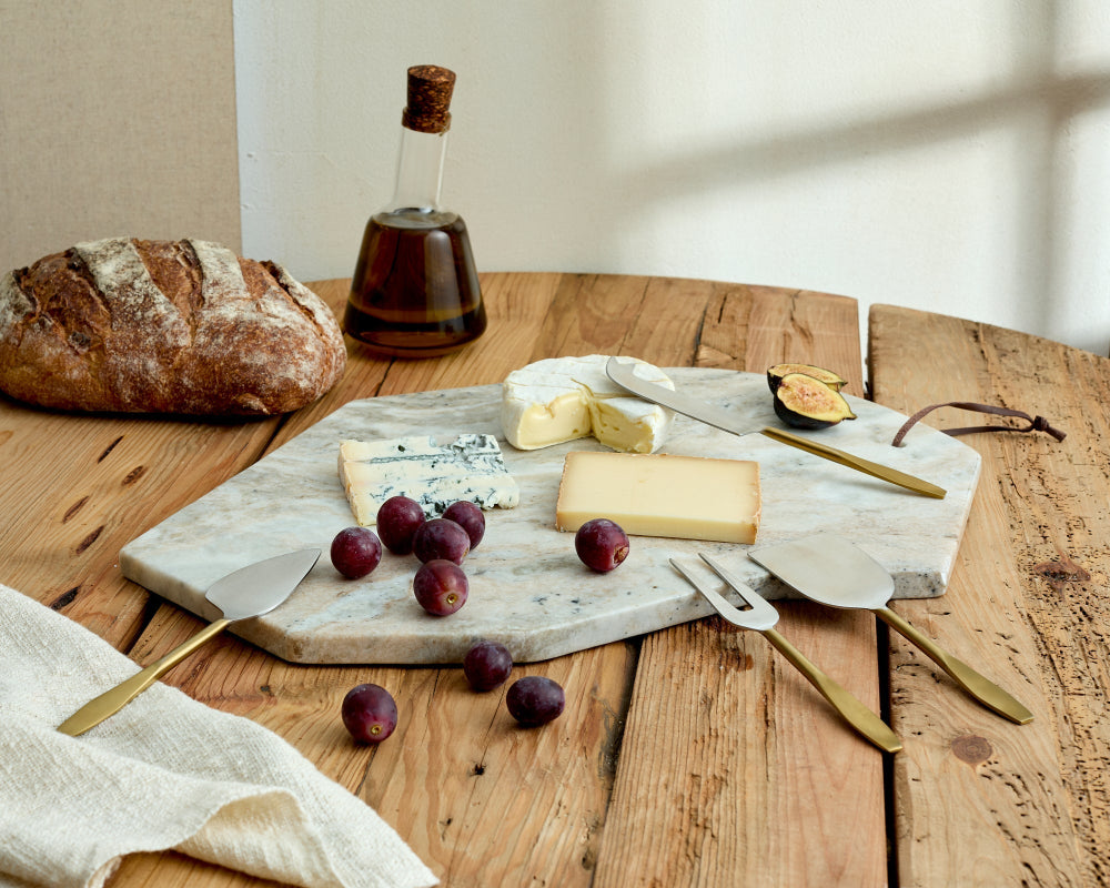 Wooden table with a cheeseboard featuring various types of cheese, fruits, and bread, with a bottle of olive oil in the background.