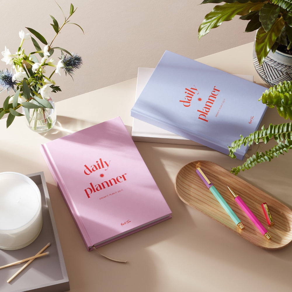 Two daily planners, one pink and one blue, on a table with pens and plants.