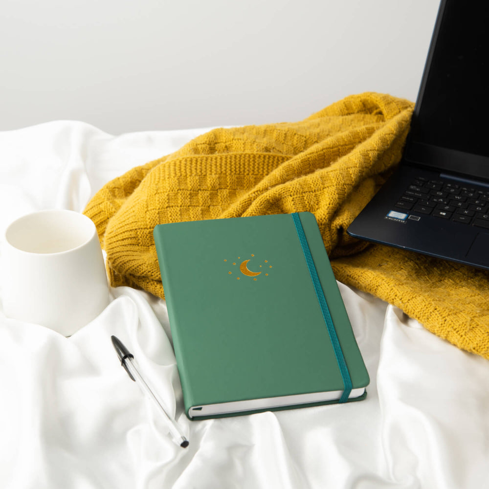 Forest Green A5 Lined notebook with a Gold debossed moon and stars, pen, yellow blanket, and laptop on white bedding.