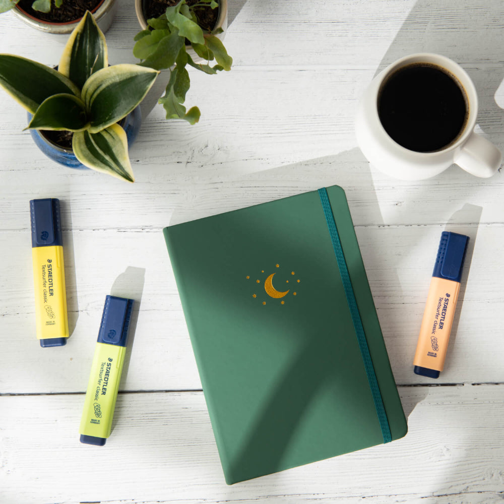 Yop and Tom Forest Green notebook with a debossed moon design on a white wooden surface with coffee, markers, and plants.