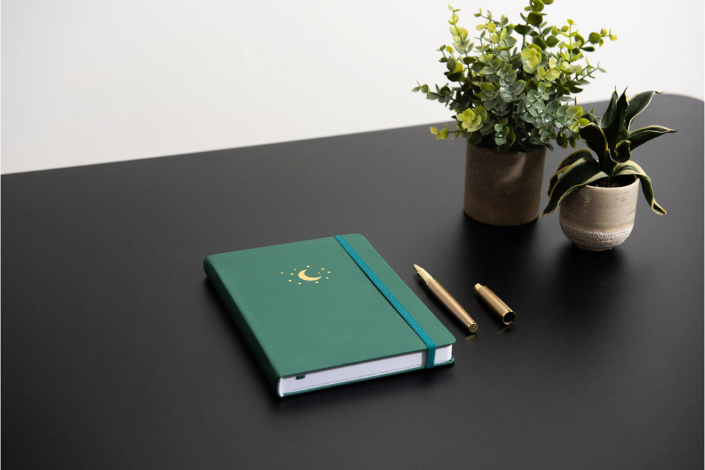 Forest Green A5 notebook on a black desk  with two potted plants and a pen.