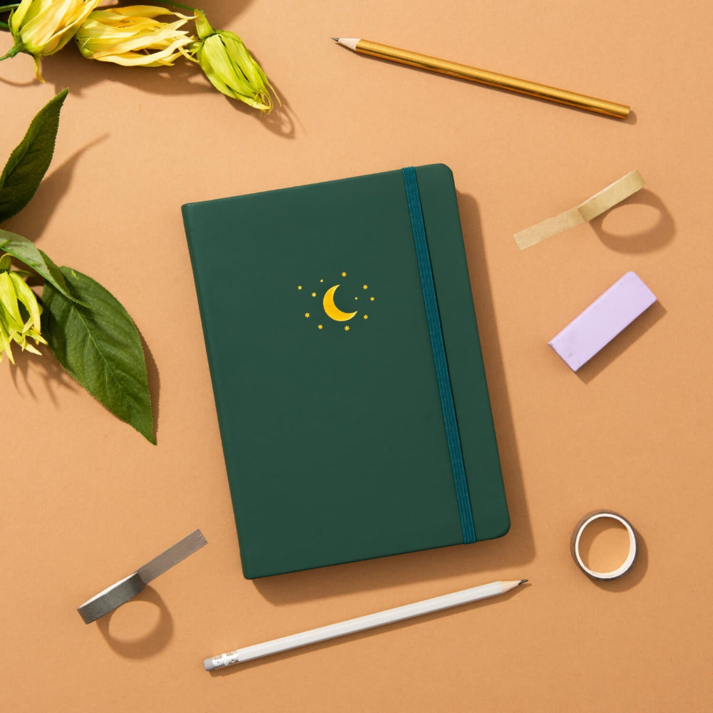 Yop and Tom Forest Green notebook with a moon and star design on a brown surface with stationery items.