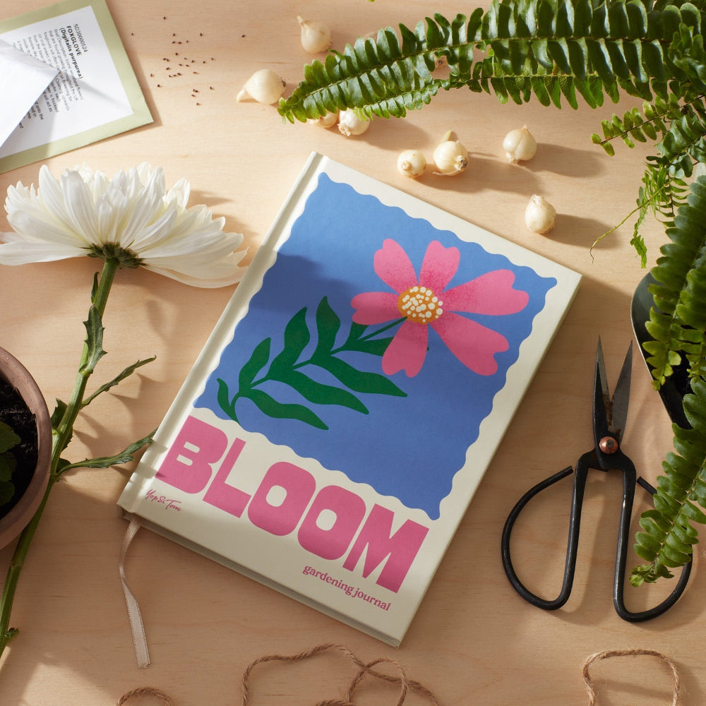 Notebook titled 'Bloom' with floral design on a wooden surface with plants and scissors.