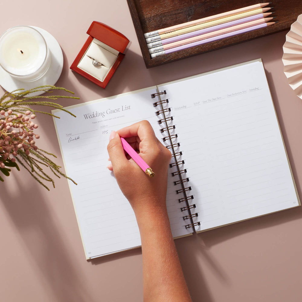Hand writing in a notebook with a pink pen, surrounded by wedding planning items on a pink surface.