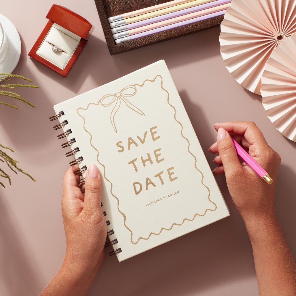 Notebook with 'Save the Date' text held by hands with a pink pen on a decorative background.
