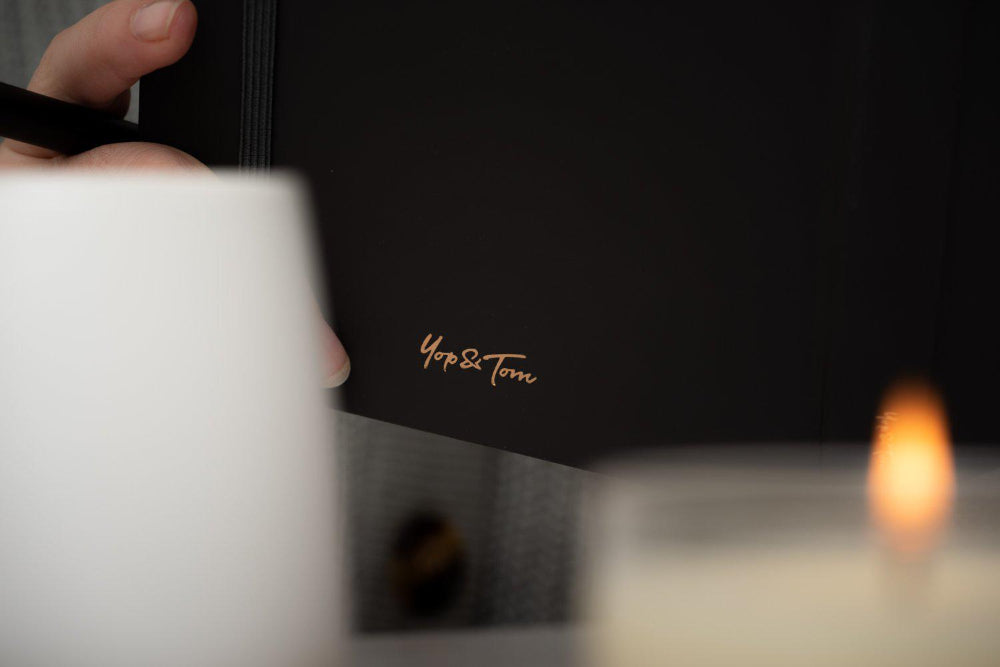 Black notebook with 'Yop & Tom' branding on a reflective surface with a blurred background.