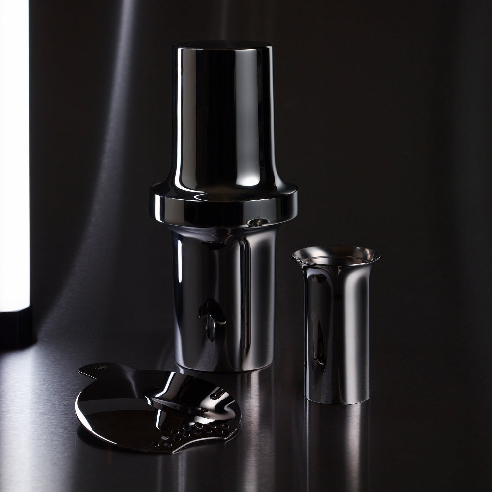 Sculptural BARBICAN stainless steel cocktail strainer in front of the matching shaker and jigger on a dark surface