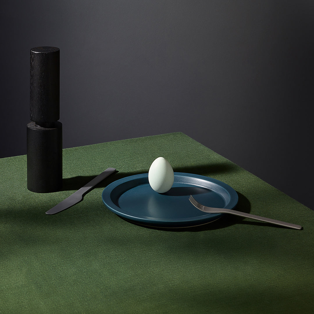 A dark blue ceramic plate with a small white egg on top, styled on a green textured tablecloth with a knife, fork, and a black pepper mill in the background.