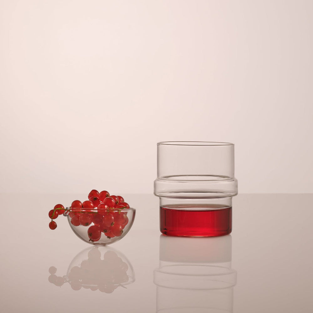 A clear glass tumbler with a stacked, ribbed design, partially filled with red liquid, shown on a reflective surface next to a small glass bowl of red berries against a warm off-white background.