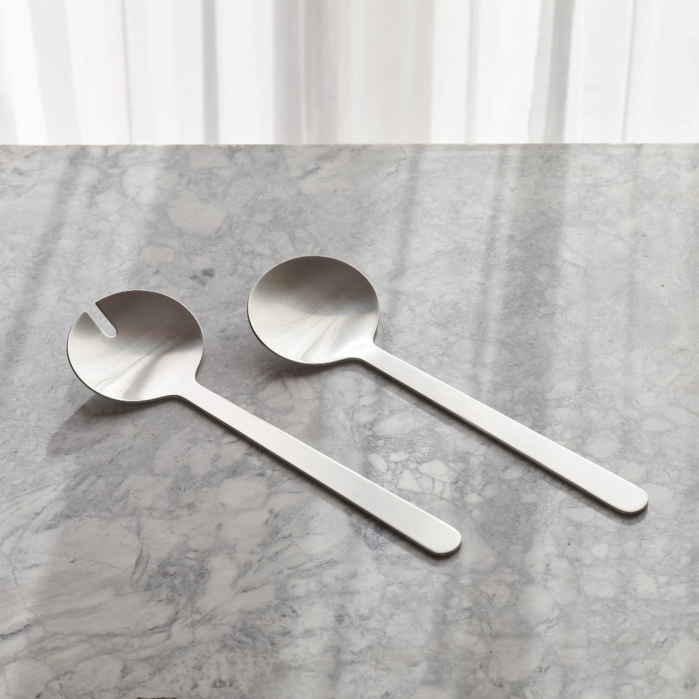 Two stainless steel serving spoons with long handles and integrated lids, resting on a gray marble surface with a soft white curtain background.